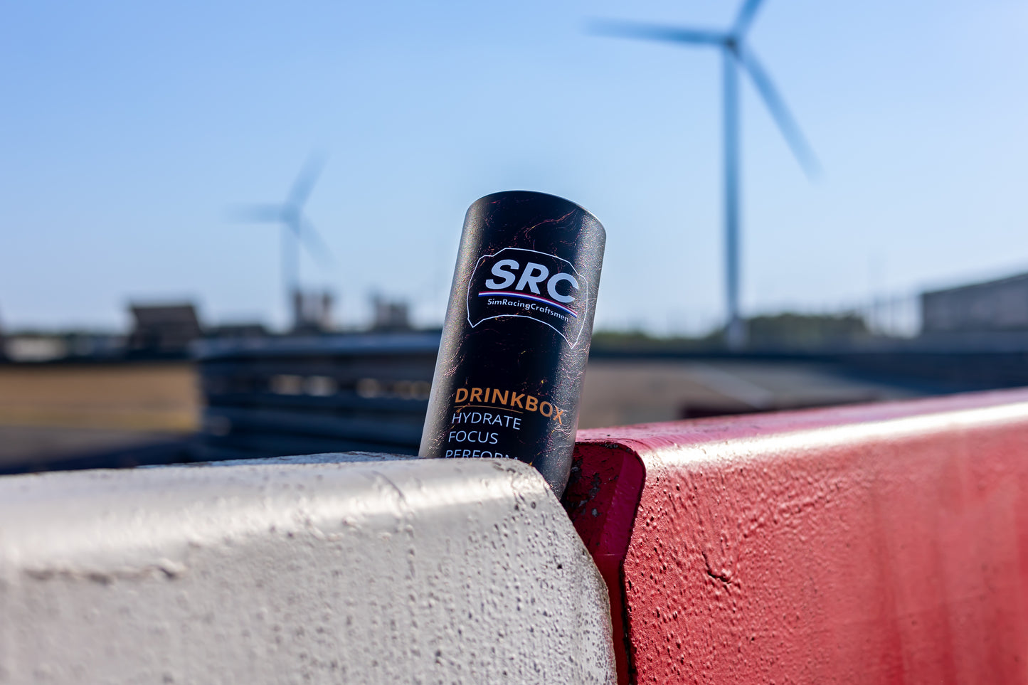 SRC race bottle leaning against a red and white barrier outdoors at a track, with wind turbines blurred in the background under a blue sky.