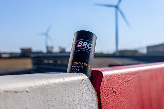 SRC race bottle leaning against a red and white barrier outdoors at a track, with wind turbines blurred in the background under a blue sky.