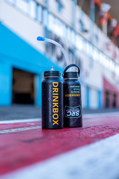 Two black Drinkbox water bottles with yellow branding, one with a clear straw and one with a black loop handle, placed on a red and white racetrack with a blurred blue building in the background.
