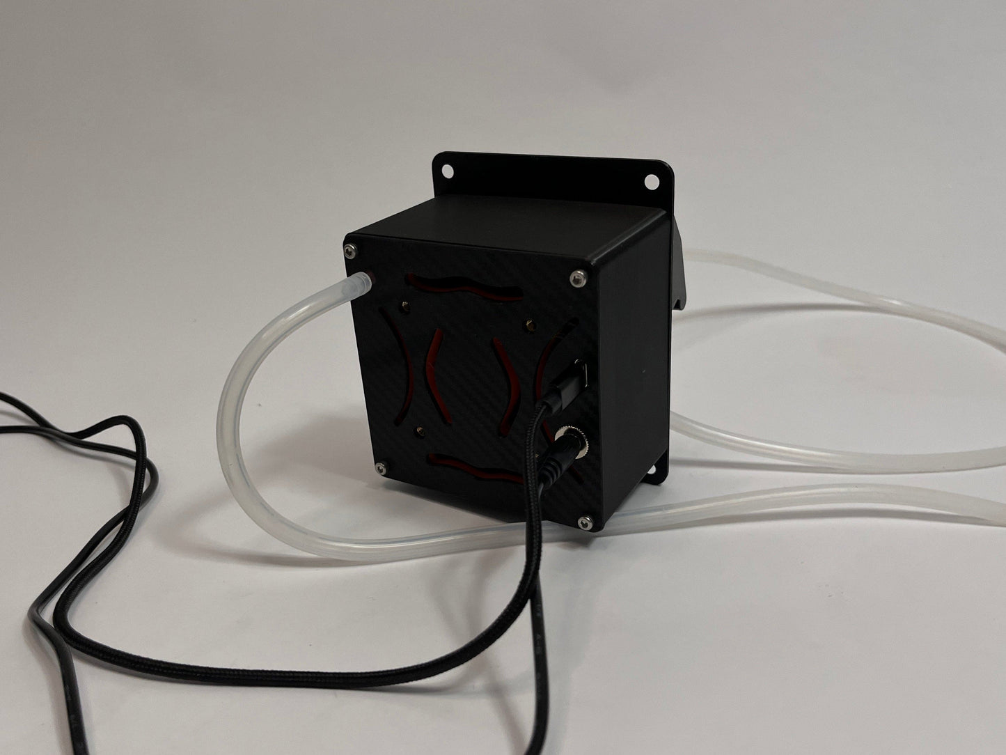 rear of the src drinkbox drinking unit for sim racing showing air cooling ducts with orange internals and a carbon fiber texture back plate. Silicone white tubing and black cables coming out of the back. On a light gray background