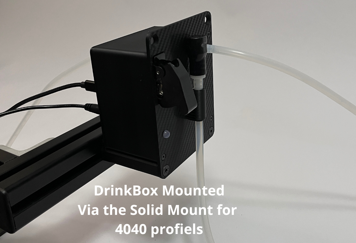 A black sim racing DrinkBox device mounted on a black solid stand with clear tubing, shown against a white background with a text overlay describing the mounting setup.