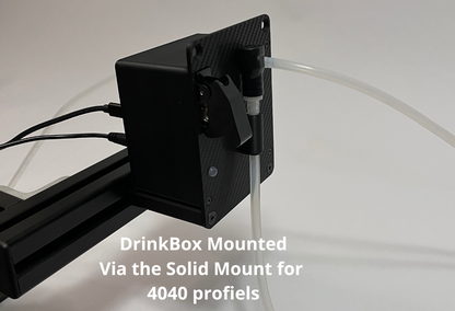 A black sim racing DrinkBox device mounted on a black solid stand with clear tubing, shown against a white background with a text overlay describing the mounting setup.