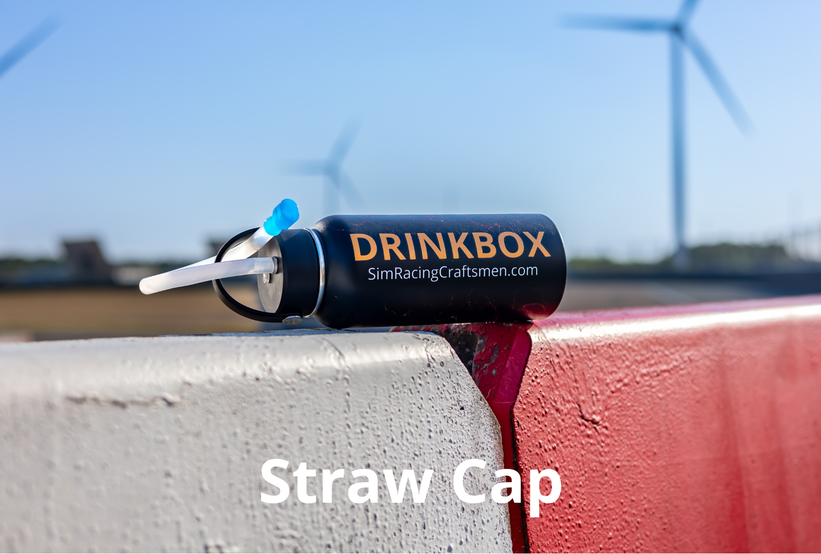 A black straw cap with orange and white branding reading 'DRINKBOX SimRacingCraftsmen.com' resting on a red and white barrier, with a blurred outdoor background of wind turbines and blue sky.